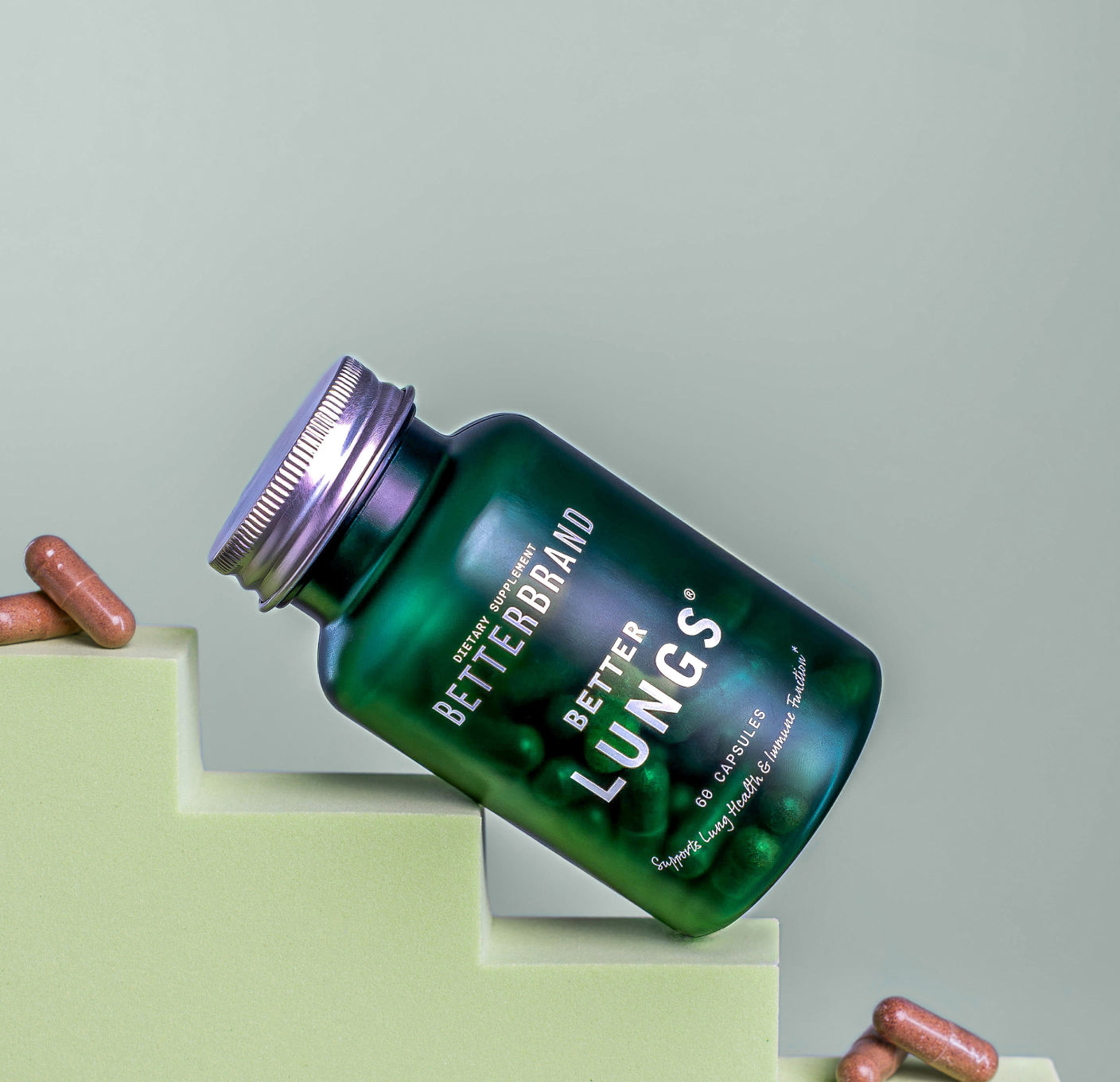 BetterBrand dietary supplement bottle labeled Better Lungs with capsules inside and some capsules outside on a green stepped surface