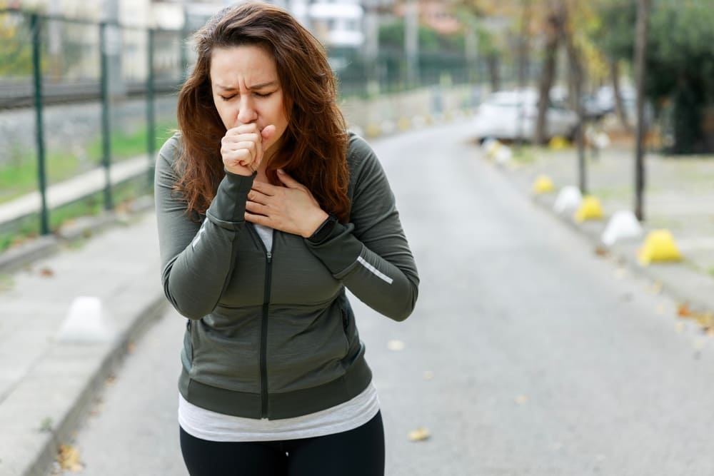 Woman outdoors coughing into her fist, holding her chest, wearing a gray jacket and black leggings.