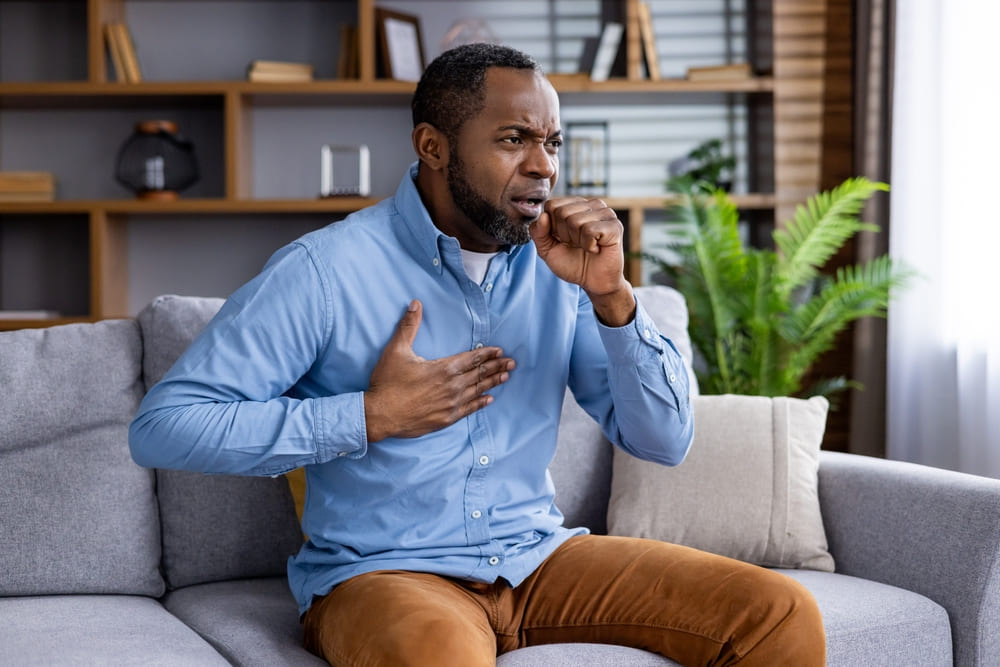 Man coughing and holding chest, sitting on a couch in a living room.