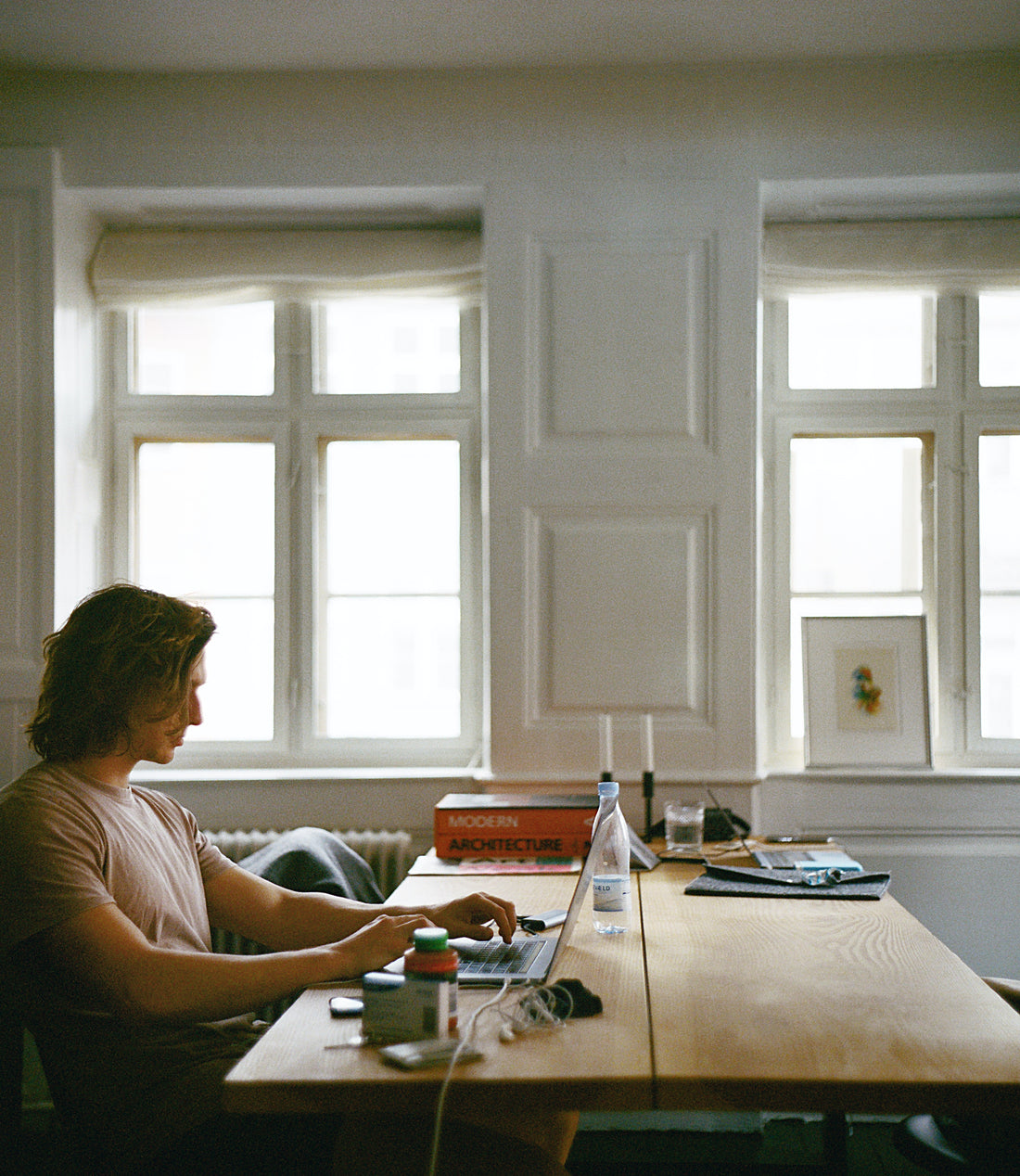 man with long hair working from home and typing on laptop at long dining room table